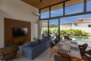 Ein modernes Wohnzimmer mit Fernseher, Sofa, Esstisch und Stühlen mit Blick auf Pool und Garten.