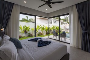 Ein modernes Schlafzimmer mit einem großen Bett, einem Deckenventilator und Blick auf Garten und Pool.