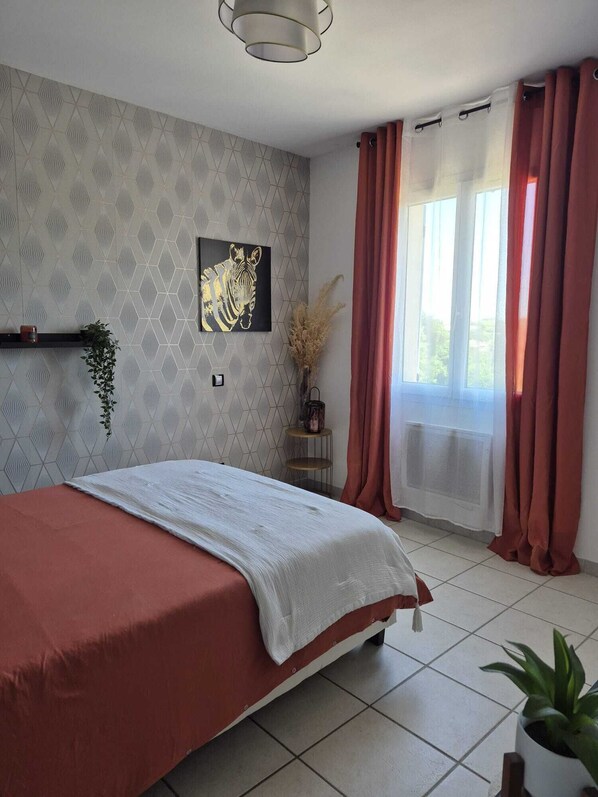 Un dormitorio con una cama, un cuadro de cebra colgado en la pared, una planta en maceta, una mesita con una vela y una ventana con cortinas.