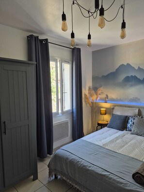 Ein Schlafzimmer mit Bett, Kleiderschrank, einem fenster mit Vorhängen, einer Wandleuchte und einem Bergpanorama.