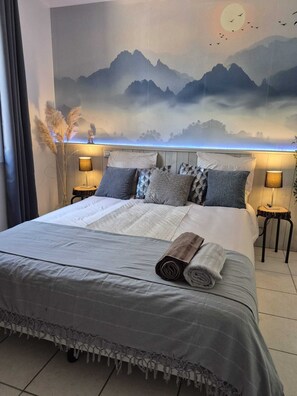 Una cama bien hecha con un mural de vistas montañosas, dos lámparas de noche y una mesita con lámpara.