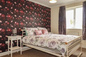 A bedroom with a floral wallpaper, a bed with a floral bedspread, a bedside table with a lamp, and a window with curtains. - Treetops (Par)