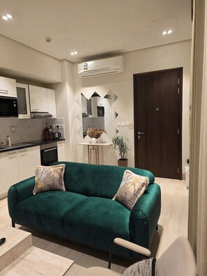 Ein modernes Wohnzimmer mit einem grünen Sofa, einem kleinen Tisch mit einer Pflanze, einer Küchenzeile mit weißen Schränken und einer dunklen Holztür.