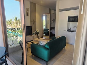 Un salon moderne avec un canapĂ© vert, un coin repas, une cuisine Ă©quipĂ©e dâun four Ă micro-ondes et une vue sur les palmiers Ă lâextĂ©rieur.