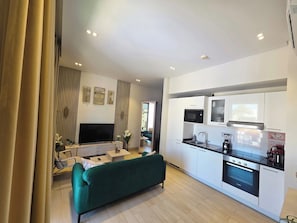 A modern living room with a green sofa, a flat-screen TV, and a kitchen with white cabinets and a built-in oven.