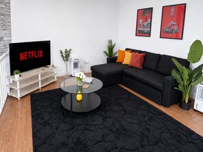 Ein Wohnzimmer mit einem schwarzen Sofa, einem runden Couchtisch mit einer Vase, einem Fernseher, der 'NETFLIX' anzeigt, und zwei gerahmten Bildern an der Wand.