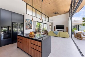 Eine moderne Küche mit Kochinsel, ein Essbereich mit Sofa und Sesseln, ein Flachbildfernseher und ein großes Fenster mit Blick auf einen Garten.
