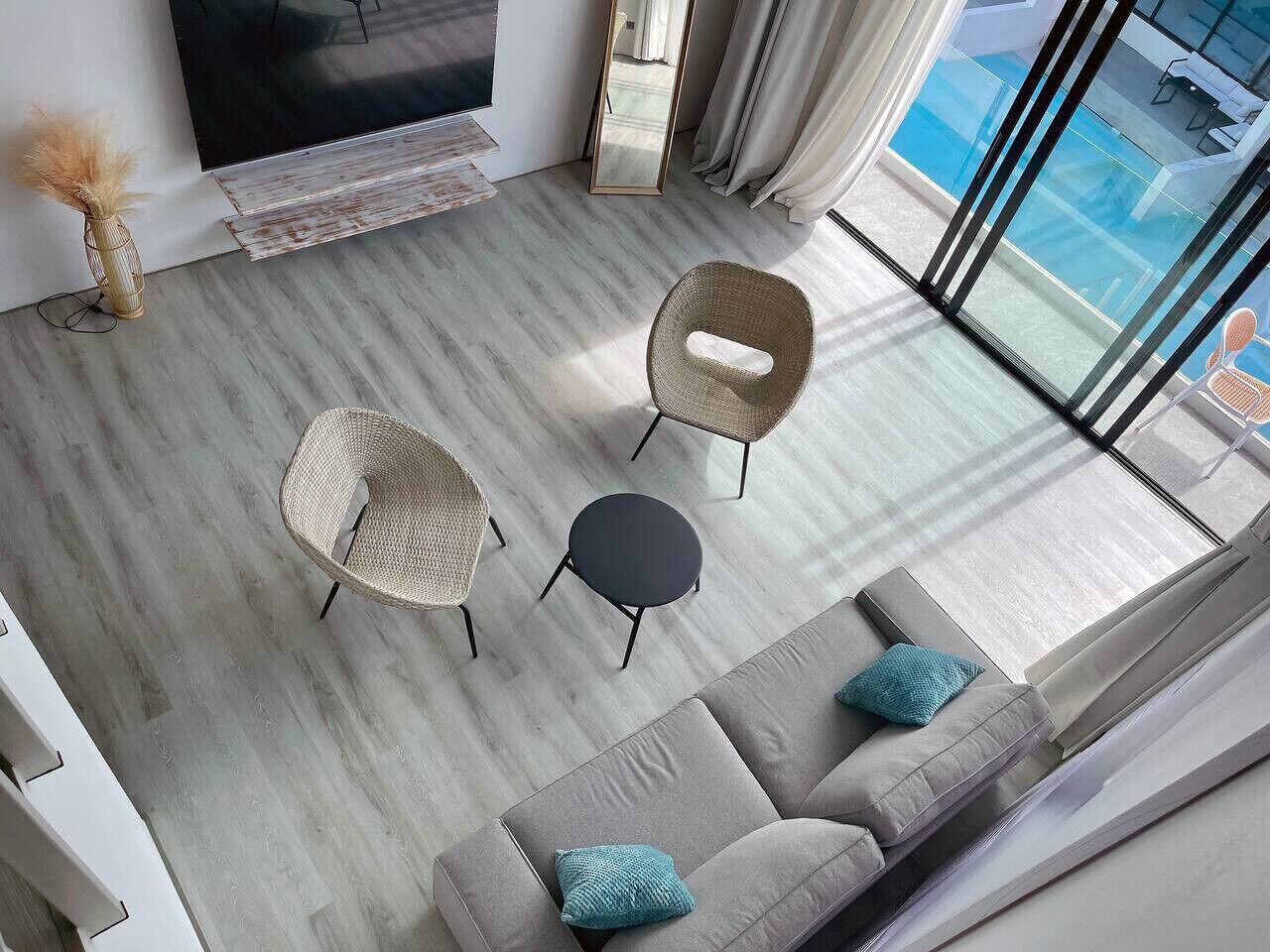 A modern living room with a sofa, two wicker chairs, a small round table, a large flat-screen TV, and a view of a swimming pool through large glass doors.