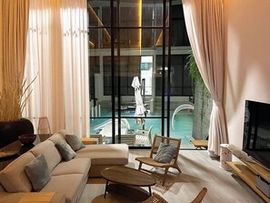 Ein modernes Wohnzimmer mit einer Couch, einem Sessel und einem Couchtisch, einem Fernseher und einem großen Fenster mit Blick auf einen Pool.
