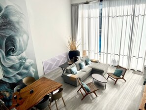 Ein modernes Wohnzimmer mit einer Eckcouch, einem Esstisch und Stühlen. An der Wand befindet sich ein großes abstraktes Wandbild, eine Vase mit Trockenpflanzen und ein Fenster mit durchsichtigen Vorhängen.