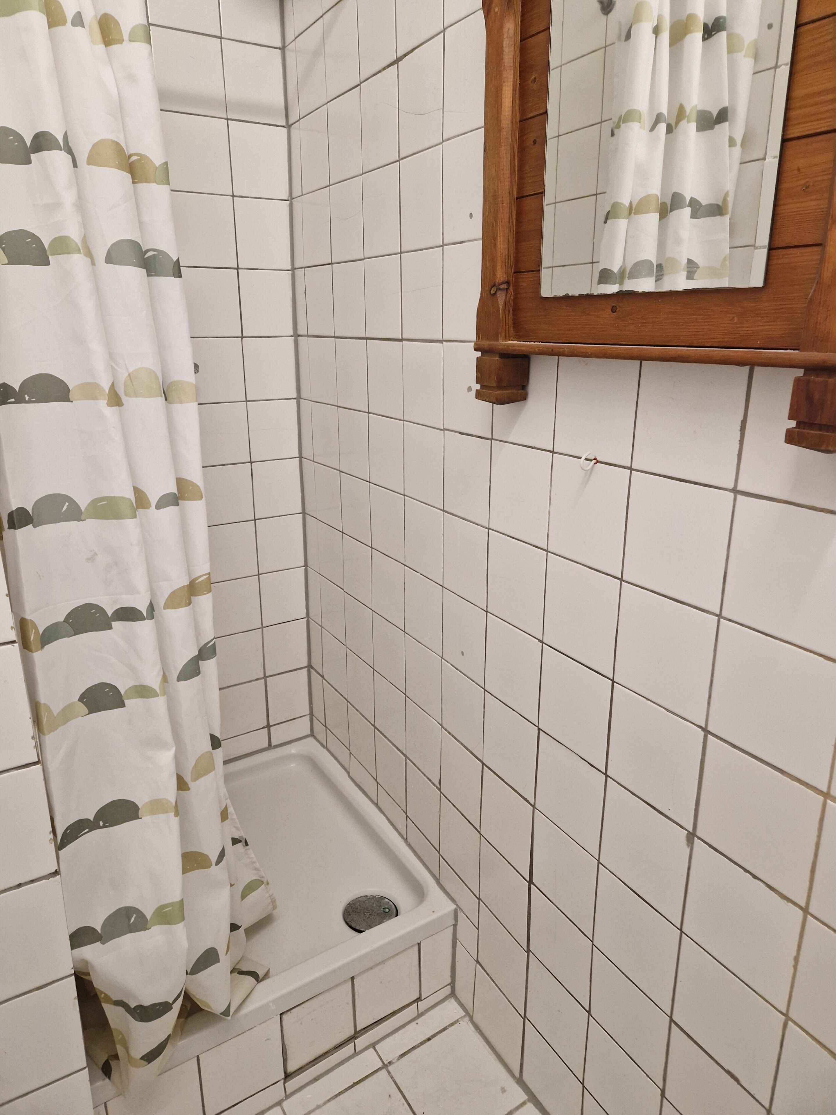A bathroom with white tiled walls, a shower curtain with a pattern, a wooden-framed mirror, and a small shower area.