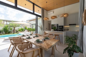 A modern kitchen with a central island, a dining area with a pool view, and a large window. - Santi Samui – 3BR Private Pool Villa (Bo Put)