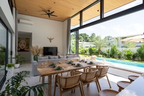 A modern living room with a dining area, large windows, a ceiling fan, and a view of a swimming pool. - Santi Samui – 3BR Private Pool Villa (Bo Put)
