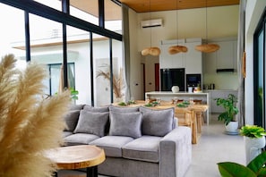 A modern living room with a grey sofa, a dining table set for four, and a kitchen in the background. - Santi Samui – 3BR Private Pool Villa (Bo Put)