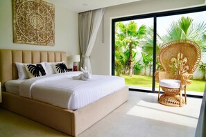 A bedroom with a large bed, a wicker chair, a view of greenery, and a decorative wall piece. - Santi Samui – 3BR Private Pool Villa (Bo Put)