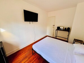 Une chambre avec un lit, un téléviseur fixé au mur, une petite table avec une bouilloire et une porte.