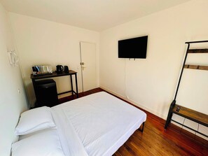 Un lit simple avec une couvre-lit blanche, un bureau avec une radio et une machine à café, un téléviseur fixé au mur et un lit superposé avec une échelle.