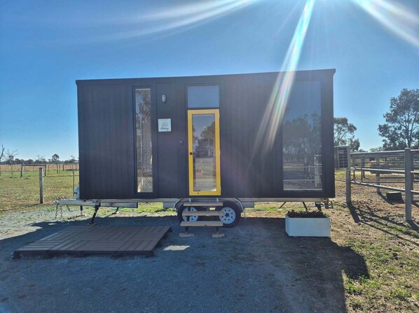 A small, dark-colored mobile home with a yellow door, a small porch, and a trailer attached for mobility. - Kookaburra Retreat by Tiny Away (Arcadia)