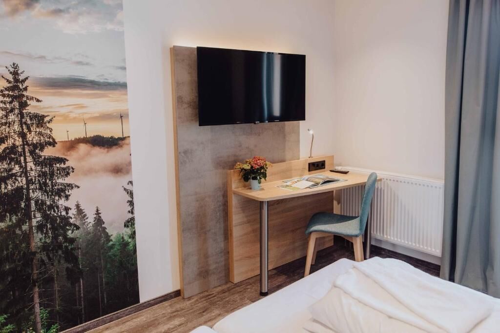 Quarto de hotel com cama, escrivaninha com cadeira, TV de tela plana na parede e vista para árvores e um pôr do sol.