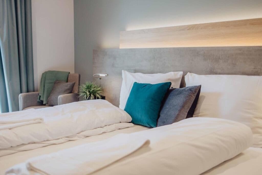 Ein ordentlich bezogenes Bett mit weißer Bettwäsche und mehreren Kissen, eine Nachttischlampe, ein Stuhl mit einem grünen Sitzkissen und eine Pflanze.