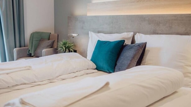 A neatly made bed with white bedding and multiple pillows, a bedside lamp, a chair with a green cushion, and a plant. - Pascals Ruhestube (Mörsdorf)