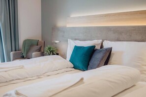 Una cama bien hecha con ropa de cama blanca y varias almohadas, una lámpara de noche, una silla con un cojín verde y una planta.