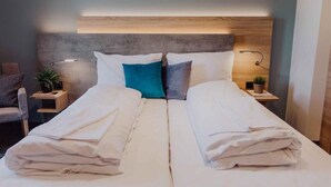 Una cama doble con ropa de cama blanca, un cabecero gris, mesitas de noche con lámparas y una planta pequeña a cada lado.