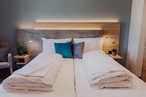 A double bed with white linens, a grey headboard, bedside tables with lamps, and a small plant on each side.