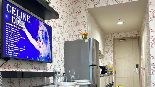 Una cocina moderna con comedor, un televisor mostrando los mayores éxitos de Celine Dion, y un cartel que dice 'MANTENGA ESTA ZONA LIMPIA'.