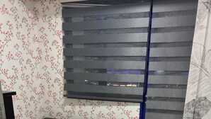 Una cama individual con una colcha de estampado de hojas en una habitación con papel tapiz floral y una ventana con persianas horizontales.
