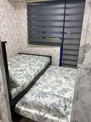 Ein Einzelbett mit Bettdecke mit Blattmuster in einem Zimmer mit floralem Wandpapier und einem Fenster mit horizontalen Jalousien.