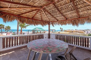 Ein überdachter Außenbereich mit rundem Tisch und Stühlen, mit Blick auf einen Strand und Gebäude.