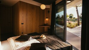 A modern bedroom with a large bed, a wooden wall, a small shelf, and a view of a landscaped garden through a glass door. - Villa Calvia Crispinilla by Villas Guide (Novigrad)