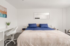 Quarto com cama, escrivaninha, cadeira, mesa de cabeceira, luminária e espelho fixado na parede.