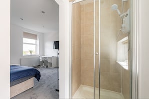 A bedroom with a bed, a desk, a chair, a shower, and a window.