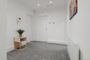 A hallway with a grey carpet, a wooden shelf with a plant, a white door, and a white radiator. - Near Anfield 3BR Apartment Free Parking (Liverpool)