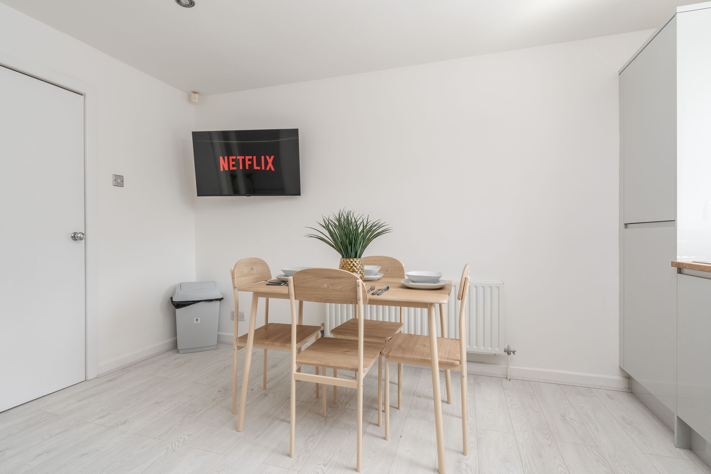 Et moderne køkken med spisebord, stole, et fjernsyn ophængt på væggen og en lille skraldespand.