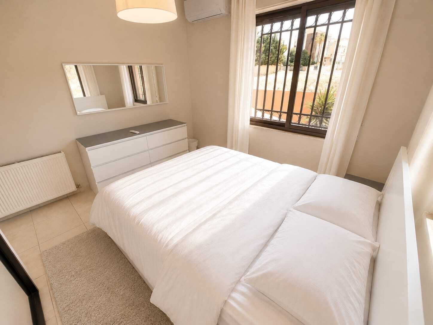 Quarto com cama, penteadeira, espelho, radiador e janela com vista para o exterior.