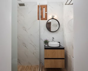 A bathroom with a glass shower enclosure, a wooden vanity with a white sink, and a round mirror. - Utopia Suites (Egina)
