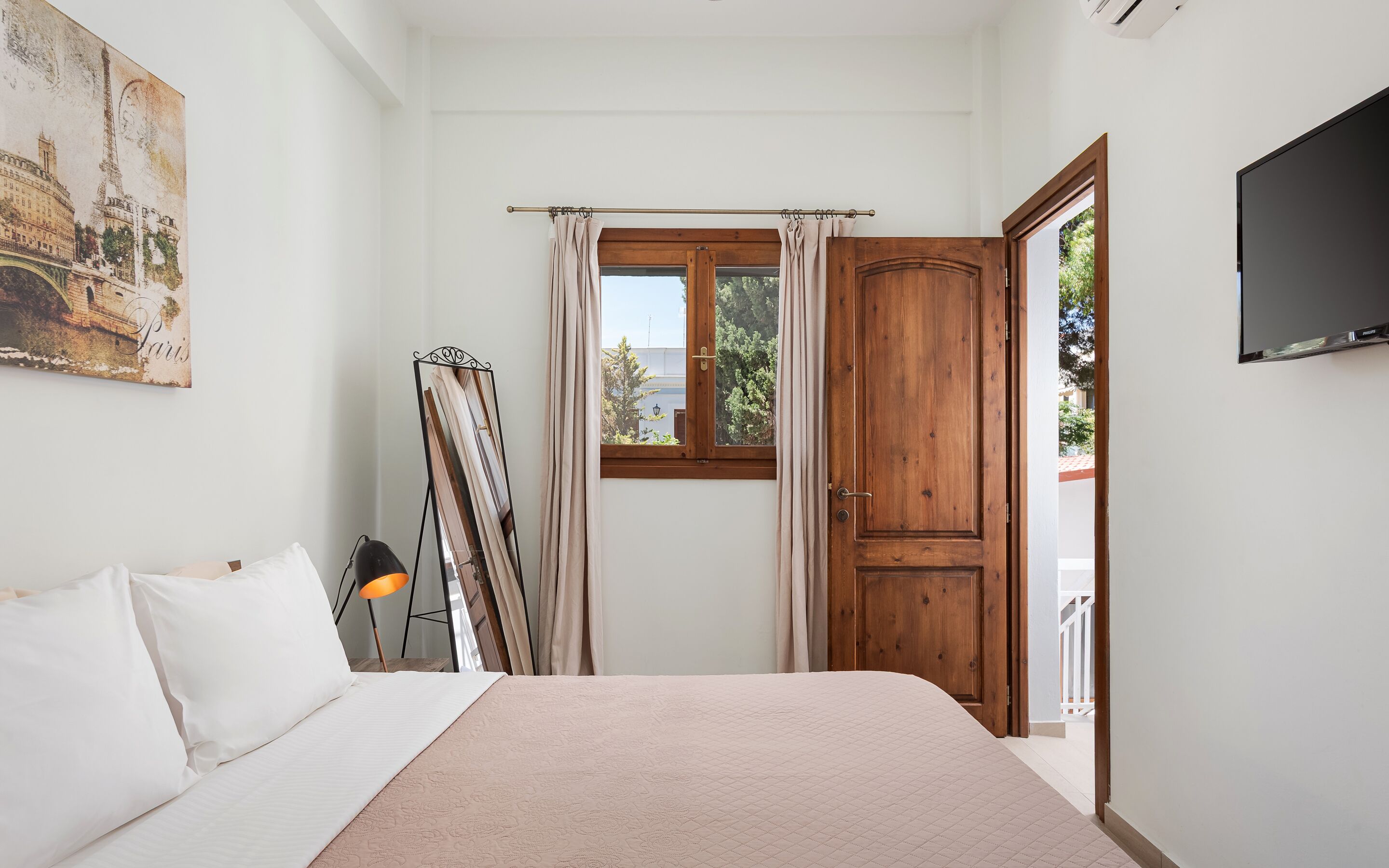 A bedroom with a bed, a painting of Paris, a wooden door, and a window with curtains.