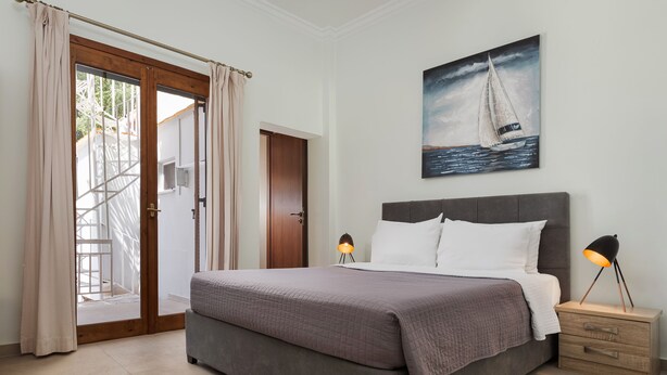 Un dormitorio con una cama, dos lámparas de noche, una mesita de noche, un cuadro de un velero y una puerta que da a un balcón.
