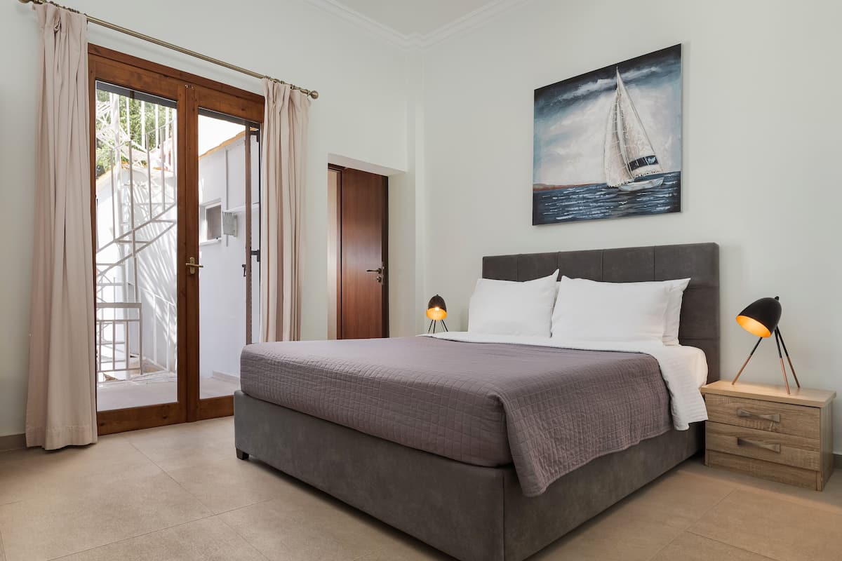 Um quarto com uma cama, duas luminárias de cabeceira, uma mesa de cabeceira, um quadro de um veleiro e uma porta que dá para uma varanda.