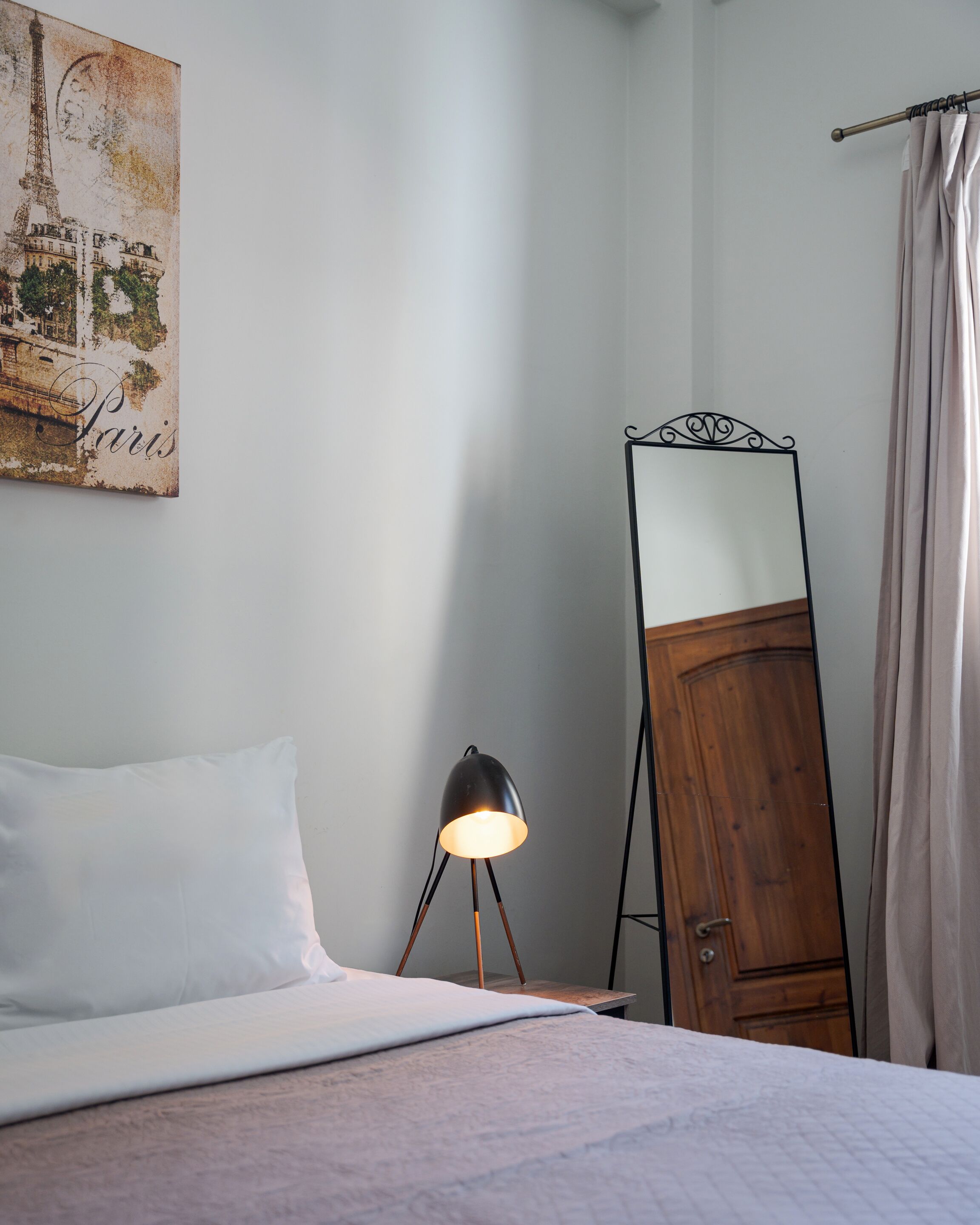 A bedroom with a bed, a standing mirror, a lamp, a painting of the Eiffel Tower, and a curtain.