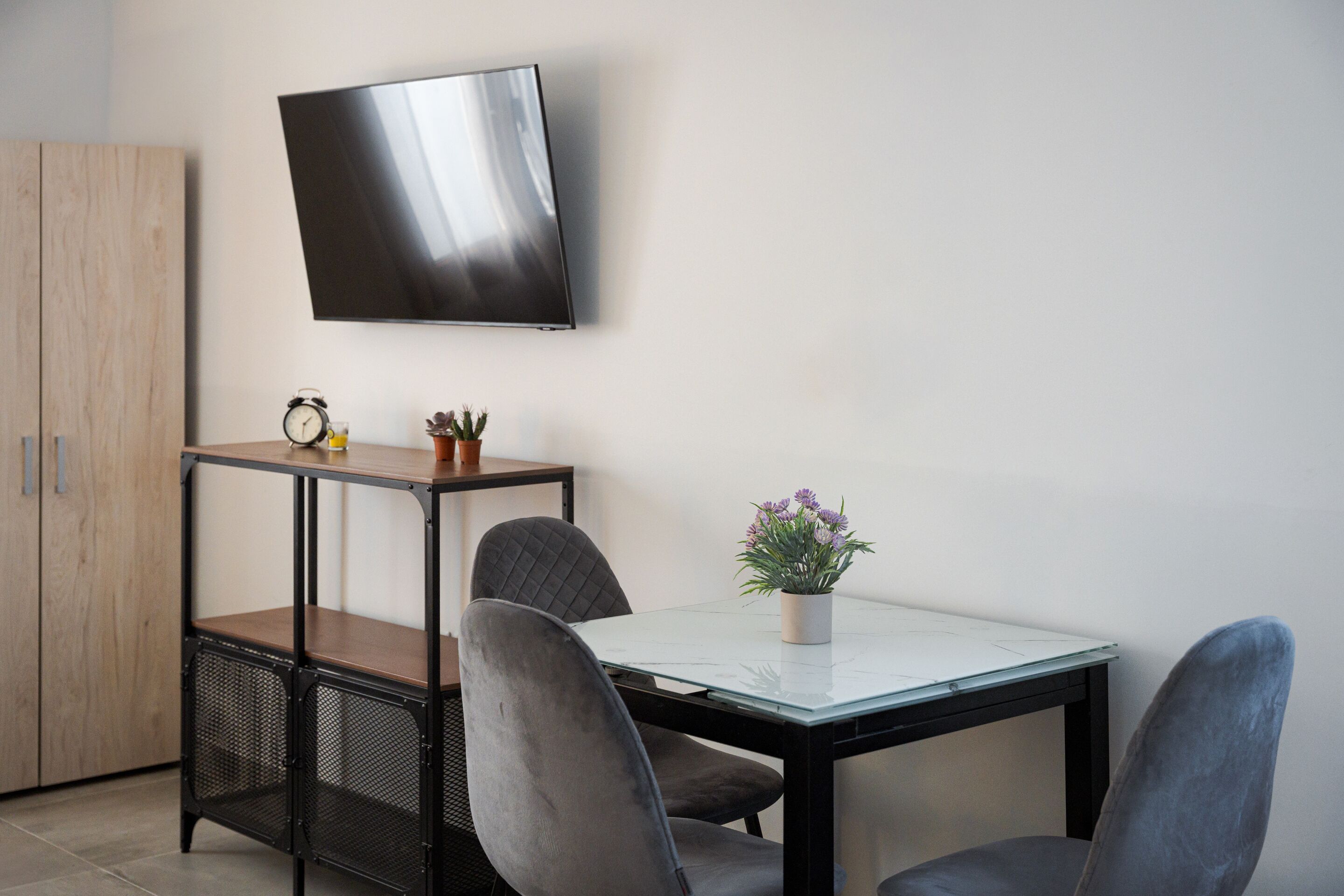 Ein modernes Zimmer mit an der Wand montiertem Fernseher, einem Holzschrank, einem kleinen Tisch mit Topfpflanze und zwei Stühlen.