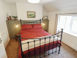A single bed with a metal frame, a red bedspread, a bedside table with a lamp, and a window with curtains. - The Monks Retreat (King's Lynn)