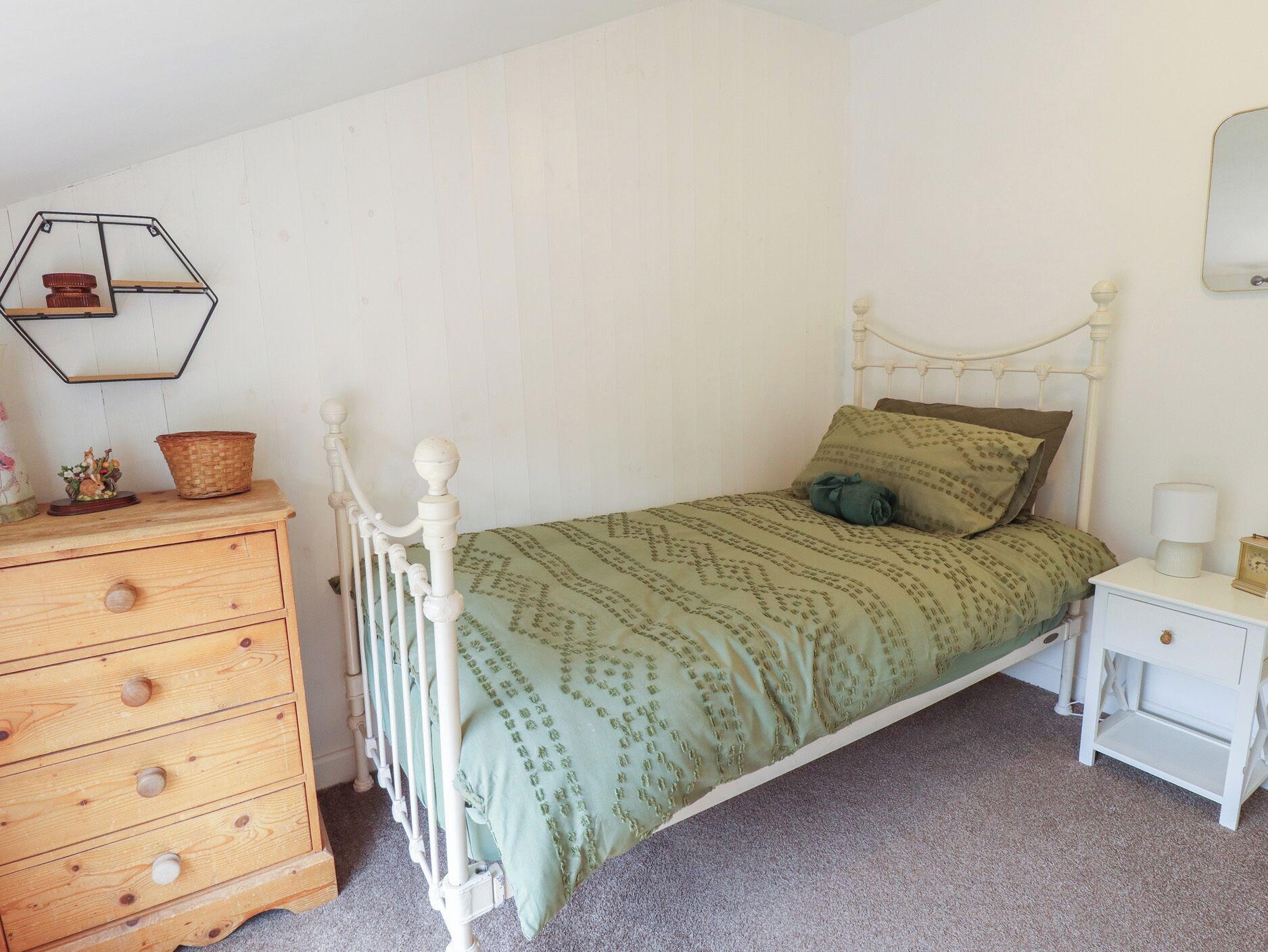 A single bed with a patterned bedspread, a wooden dresser with drawers, a white nightstand, and a wall-mounted mirror.