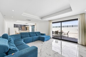 Ein modernes Wohnzimmer mit einem blauen Ecksofa, einem Essbereich mit Tisch und Stühlen und einer großen Schiebeglastür, die auf eine Außenterrasse führt.