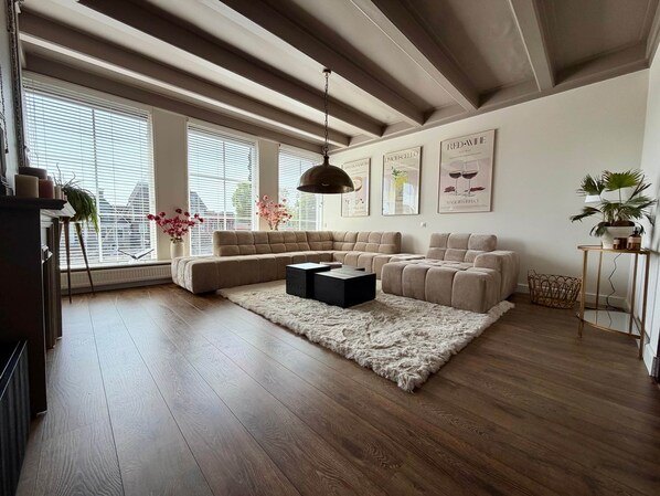 Ein modernes Wohnzimmer mit einer Eckcouch, einem Couchtisch und einem flauschigen Teppich. Große Fenster mit Jalousien, ein Deckenventilator und Wandbilder sind ebenfalls vorhanden.