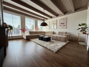 A modern living room with a sectional sofa, a coffee table, and a fluffy rug. There are large windows with blinds, a ceiling fan, and wall art.