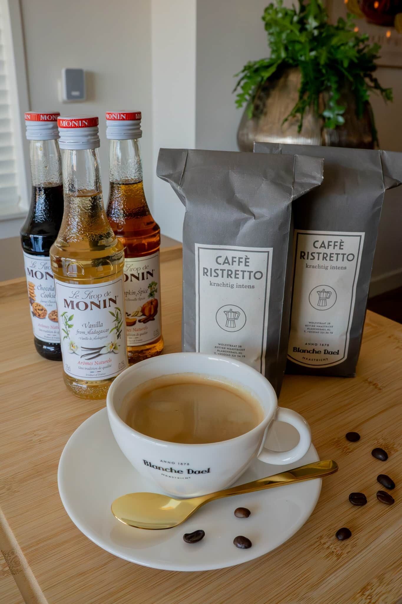 A table with a cup of coffee, three bottles of MONIN syrup, and two bags of Caffe Ristretto.